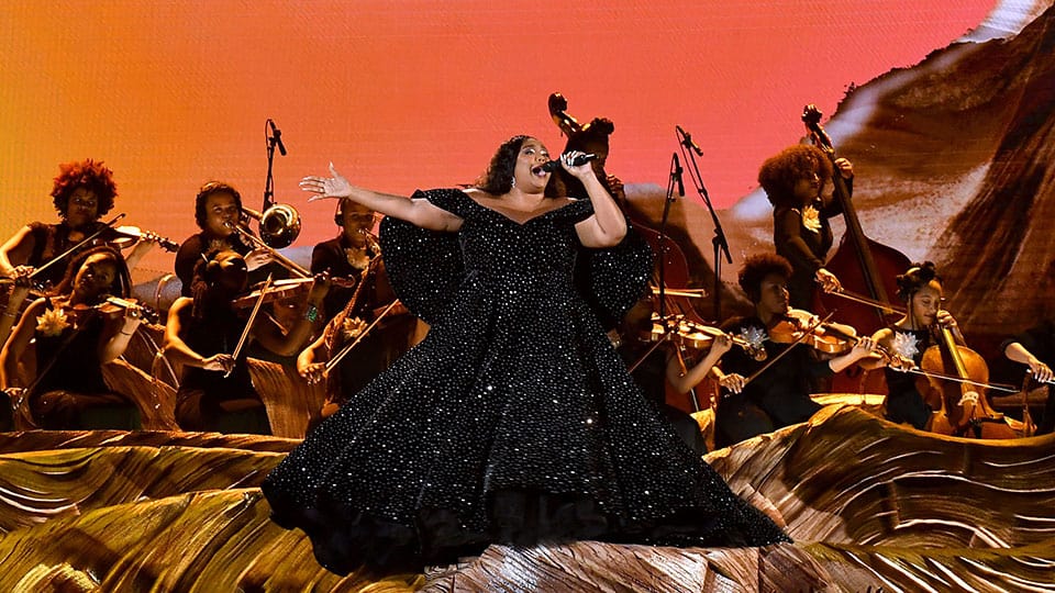 Singer Lizzo wears a black evening dress while singing in front of an orchestra of musicians at her 2020 performance at the Grammys.